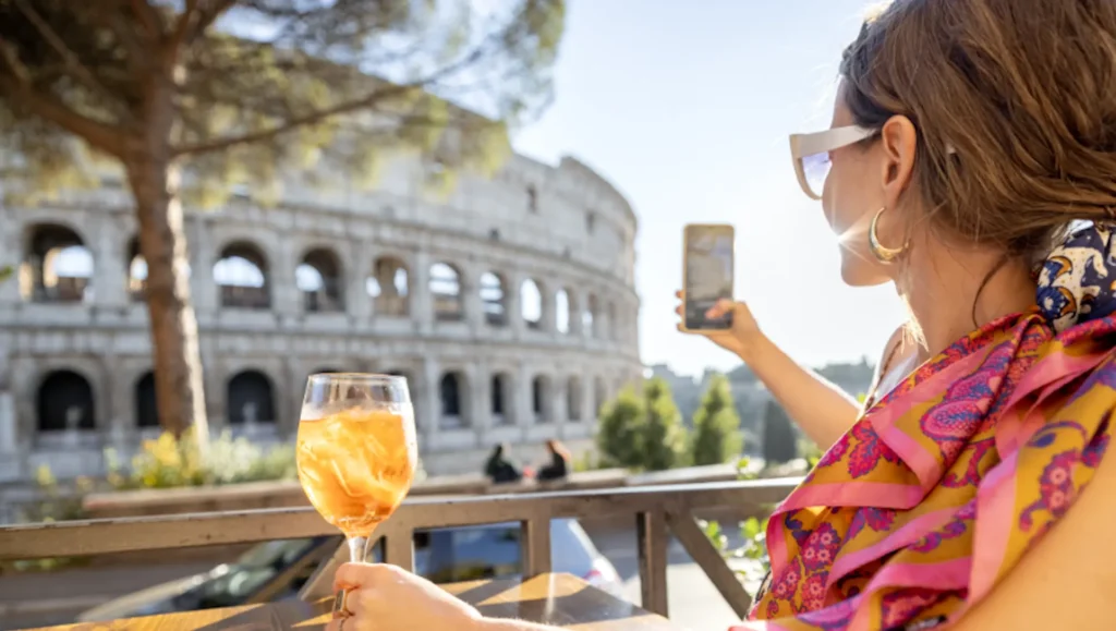 A woman with an Aperol spritz, taking a photo of the Colosseum in Rome. 